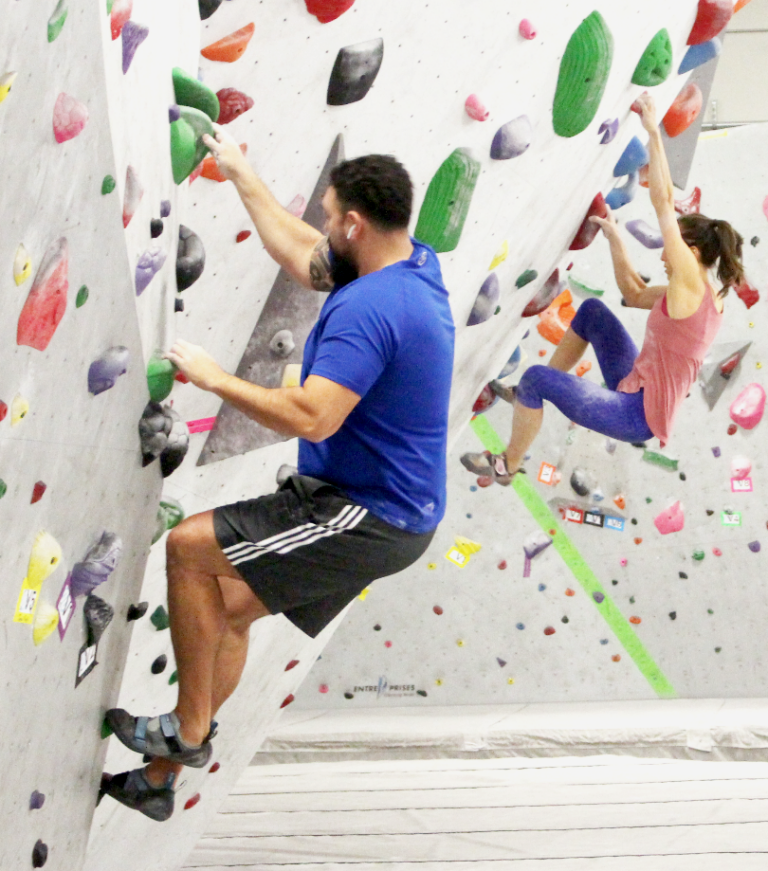 A man on shorts and tee shirt and a woman on jean and top climb a bouldering wall