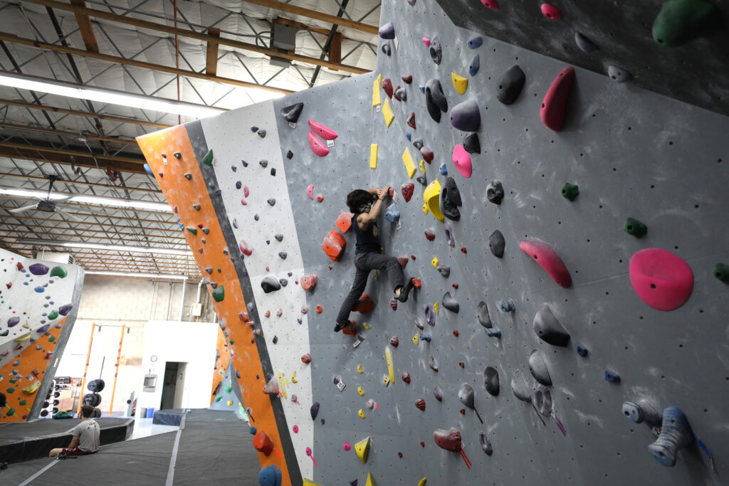 A man in black sleeveless top and black pants bouldering a gray, white and orange colored wall at The Pad gym