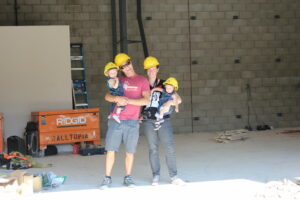 A man carrying a baby and a woman standing next to him with a camera hanging around her neck while also carrying a baby, all of them wearing yellow helmets