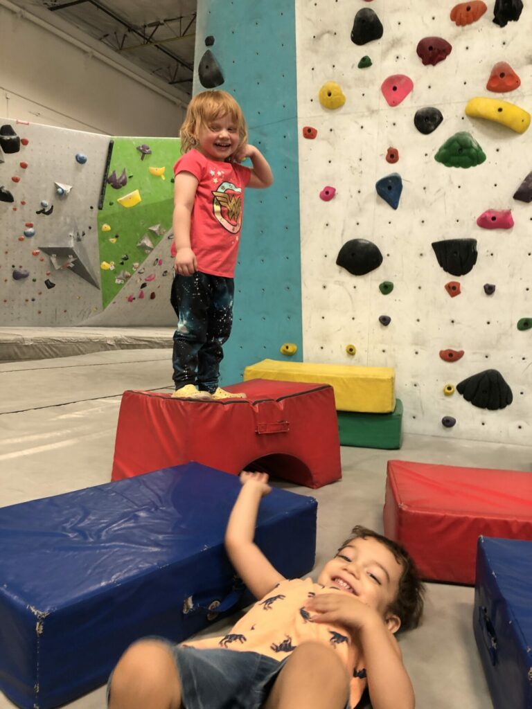 Two bubbly toddlers at the Henderson gym with one lying down and one standing on a foam
