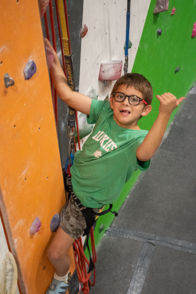 Kid with glasses and green shirt gives a thumbs up on the climbing wall